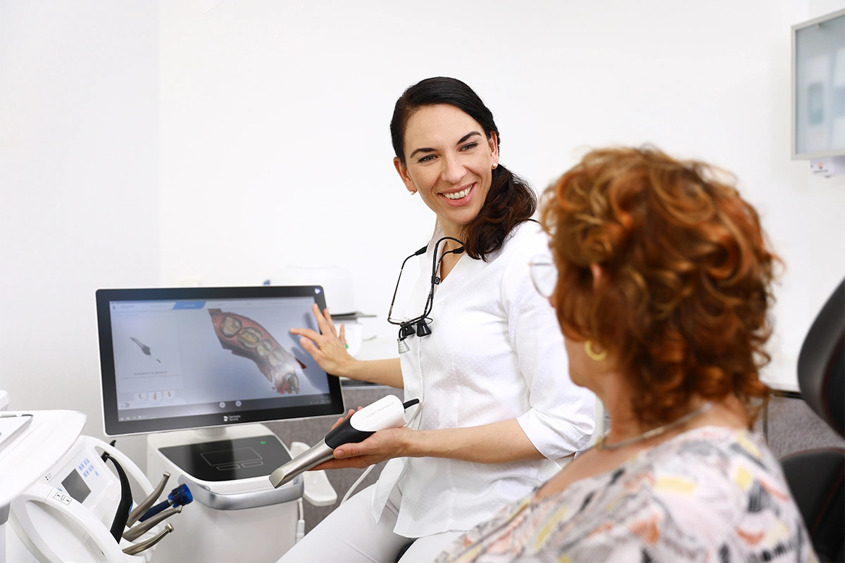 Beratungsgespräch im Behandlungsraum: Zahnärztin erklärt einer Patientin am Bildschirm eine digitale 3D-Darstellung des Gebisses und zeigt moderne Diagnostiktechnik.