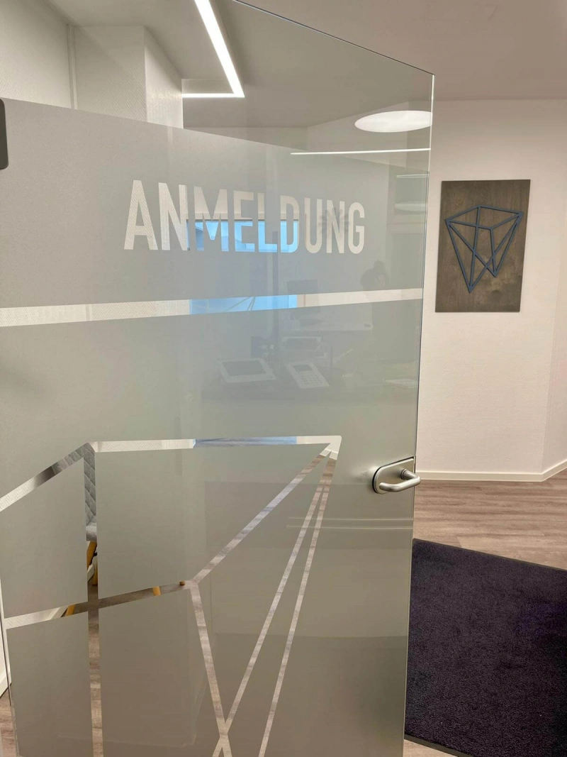 Glastür mit der Aufschrift „ANMELDUNG“ und modernem geometrischem Design – Eingang zum Empfangsbereich in hell und stilvoll gestalteten Praxisräumen.